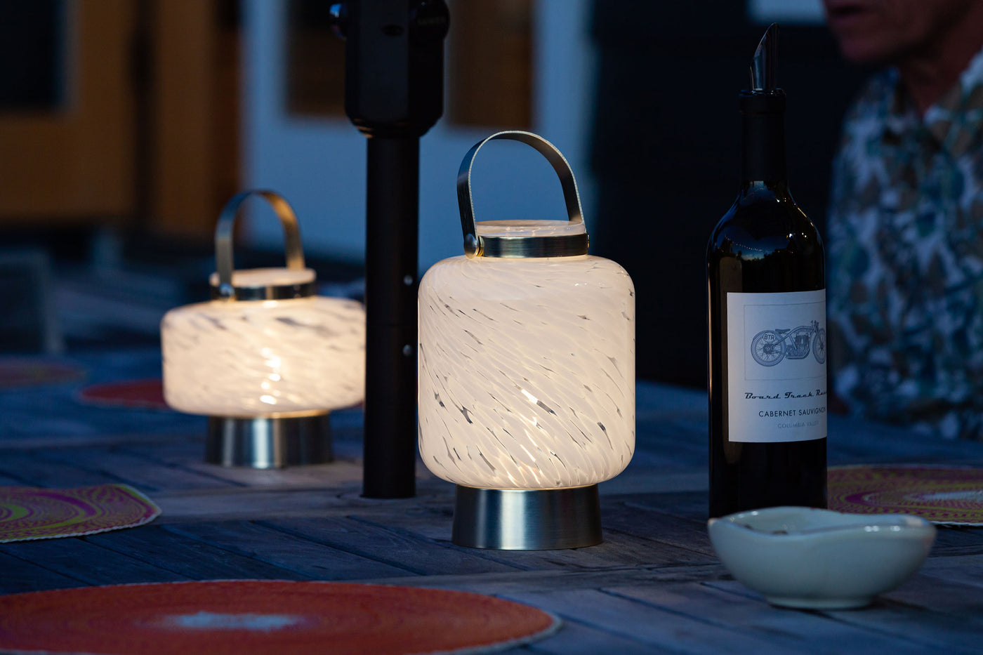 Two Illuminated LightKeeper Lanterns on an outdoor table near a bottle of wine.