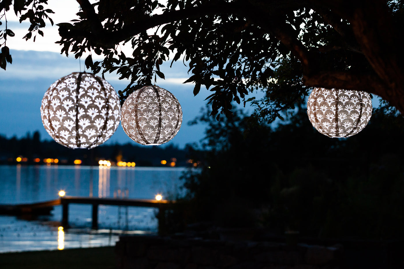Three illuminated Soji Stella Deco lanterns hang in a lakeside tree.